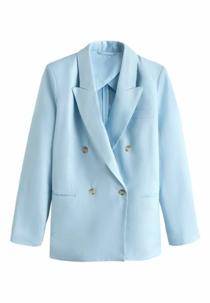 Kvinde i lyseblåt jakkesæt og sorte sandaler står på en hvid buet trappe mod en klar blå himmel.