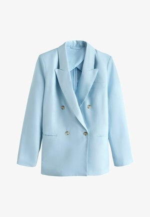 Kvinde i lyseblåt jakkesæt og sorte sandaler står på en hvid buet trappe mod en klar blå himmel.