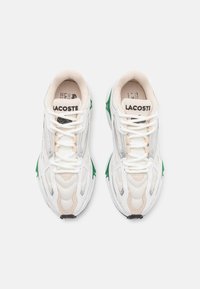 Lacoste sneakers feature a white mesh upper with beige and silver accents, green details on the sole, and white laces.