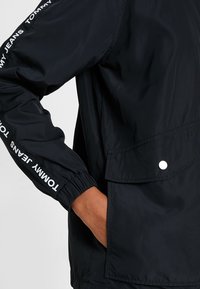 Černá lehká bunda s elastickými manžetami, boční kapsou a detaily loga "Tommy Jeans" na rukávu. Hladký, voděodolný materiál.