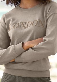 Béžová mikina s vyšívaným nápisem "LONDON" v světle hnědé barvě. Má kulatý výstřih a žebrované manžety a lem. Jemná textura látky.