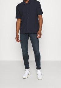 Camisa de botones de manga corta en color navy, hecha de tejido suave, combinada con jeans oscuros de corte slim y zapatillas blancas con detalles en negro.