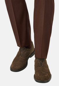 Scarpe brogue in suede marrone con dettagli perforati, abbinate a pantaloni marroni su misura che evidenziano una piega ben definita.