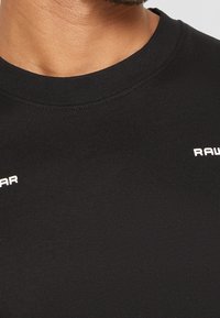 Gros plan d'une personne portant un t-shirt noir à col rond avec un texte partiel blanc "RAW" et "AR" visible sur la zone de la poitrine.
