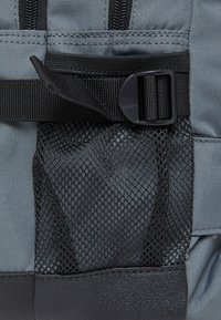 Sivi ruksak s teksturiranim materijalom, crnom mrežastom bočnom džepom, podesivim remenom s plastičnom kopčom i ojačanim dnom.