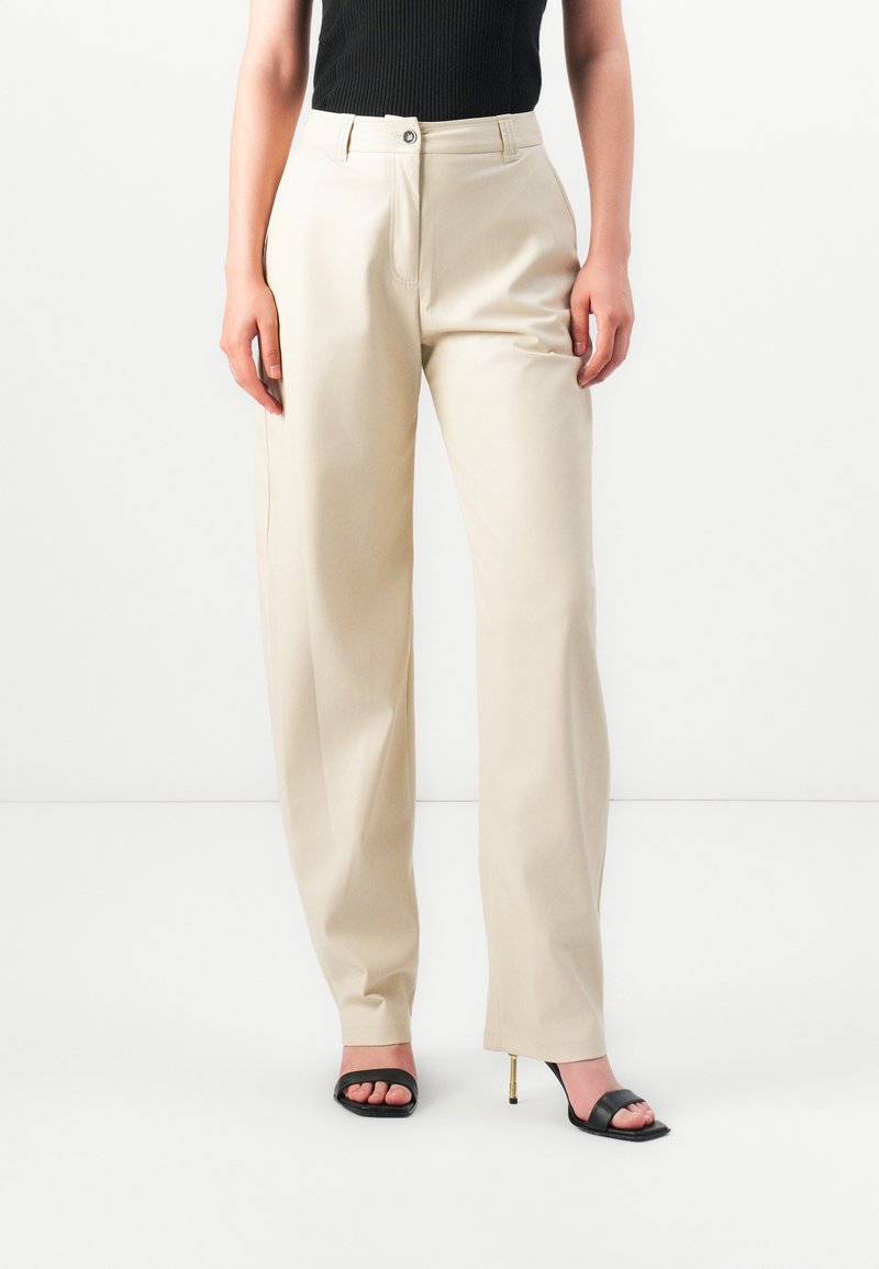 Donna che indossa pantaloni dritti color crema, top senza maniche nero e sandali con tacco aperto neri su sfondo bianco.