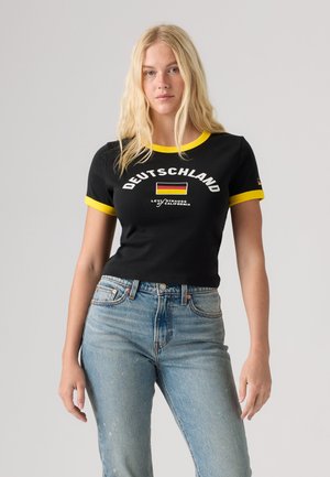 Žena má na sebe čierne tričko so žltým lemom, textom "Deutschland", nemeckou vlajkou a logom Levi's, v kombinácii so svetlomodrými džínsami na sivom pozadí.