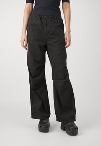 Pantalons larges noirs en tissu léger, avec une taille élastique et un minimum de coutures. Associés à des sabots noirs.