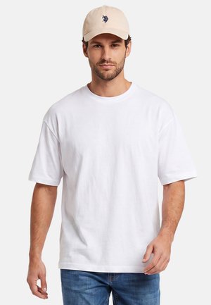 Man wearing a beige cap, plain white oversized t-shirt, and blue jeans, standing against a plain light background.