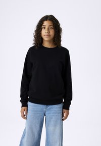 Schwarzer Sweatshirt mit langen Ärmeln und gerippten Bündchen, kombiniert mit hellblauen weitem Jeans, vor einem schlichten weißen Hintergrund.