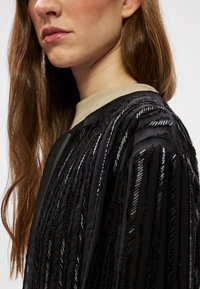 Μαύρο διαφανές ύφασμα με κάθετες υφασμάτινες ρίγες και παγιέτες. Το μ�οντέλο φοράει μια μπεζ κολάρο από κάτω. Τα μαλλιά είναι μακριά και κυματιστά.