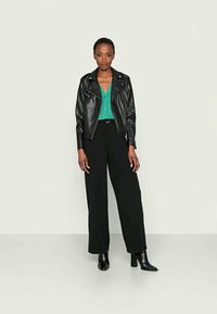 Jaqueta de motoqueiro em couro preto sobre uma blusa verde, combinada com calças largas pretas de cintura alta e botas pretas de cano curto. Design simples e estruturado.