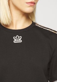 Lähedalt vaadatuna on isik, kes kannab musta Adidas t-särki, mille õlgadel on leopardimustrilised triibud ning Adidas logo rinnal.