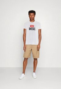 Vyras stovi prieš paprastą sieną, vilkintis baltu "Diesel" logotipą turinčia marškinėliais, bežė šortais ir baltais sportbačiais, žiūri į priekį, rankos atpalaiduotos.