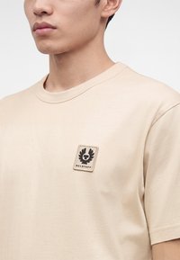 Млад мъж, носещ бежова тениска с къс ръкав и квадратен логотип на Belstaff на лявата страна на гърдите, на едноцветен фон.