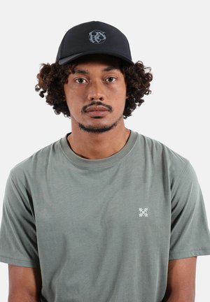 Homme aux cheveux bouclés portant un t-shirt uni vert olive et une casquette noire avec un logo abstrait circulaire, faisant face à l'avant sur un fond blanc.