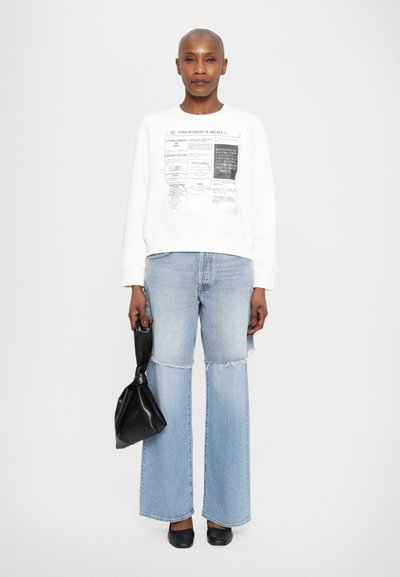 White sweatshirt with graphic text, light blue wide-leg jeans with frayed hem, and black bag held in left hand. Black shoes complete the look.