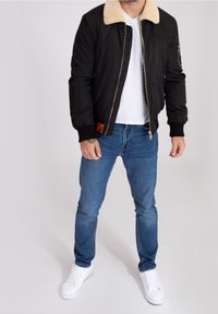 Svart bomberjacka med en krämfärgad fuskpälskrage, dragkedja fram, ribbade muddar, bärs över en vit T-shirt och blå jeans. Vita sneakers fulländar looken.