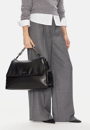 Osoba trzymająca dużą czarną skórzaną torebkę na łańcuszku, ubrana w szare szerokie spodnie, szary sweter i białą koszulę.