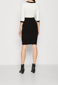 Kvinna står med ryggen mot kameran, iklädd en svart blyertskjol, vit topp med svart kant, och svarta stövletter på en enkel ljus bakgrund.