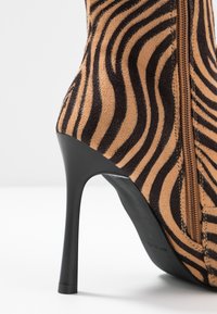 Botín de tacón alto en ante con estampado de cebra en tonos marrón y negro, que cuenta con una cremallera lateral y un elegante tacón negro. El patrón texturizado añade profundidad.