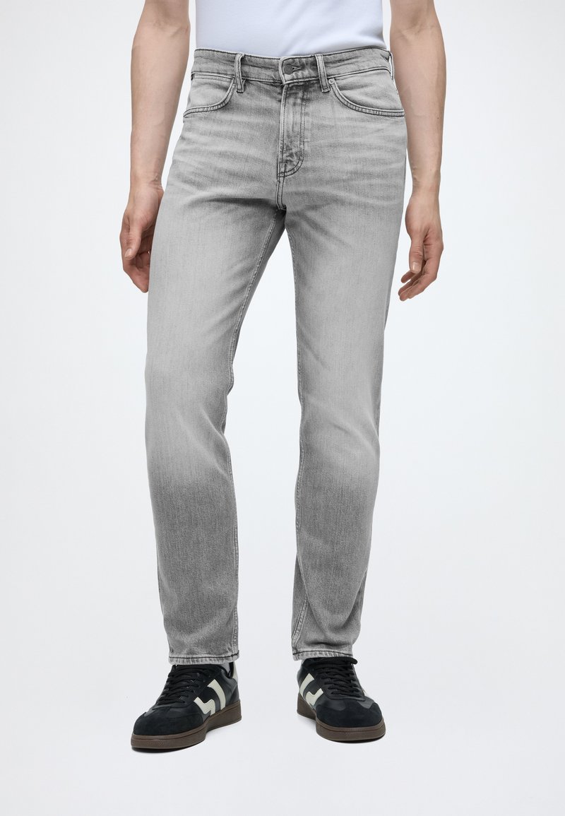 Personne portant un jean slim gris clair et des baskets noires avec des touches blanches, debout devant un fond blanc uni.