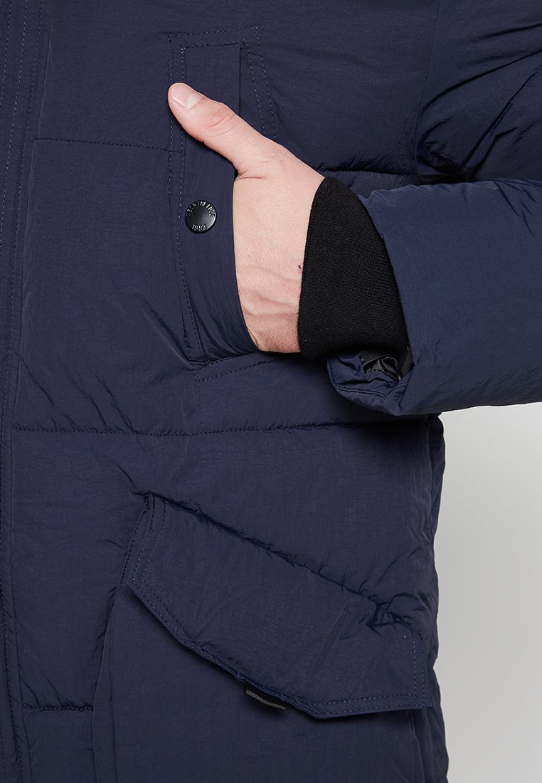 Veste matelassée bleu marine avec une surface texturée, dotée d'une poche poitrine à bouton et d'une poche latérale en biais. Détails des poignets en noir visibles.