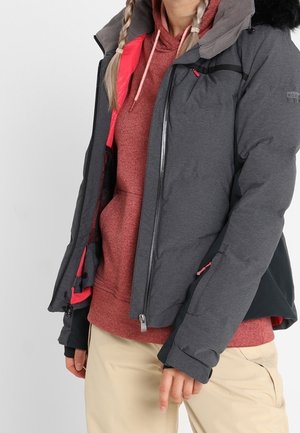 Personne portant une veste d'hiver grise avec des détails noirs et roses sur un sweat à capuche rouge et un pantalon beige, debout devant un fond uni.