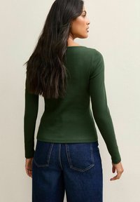 Maglia verde a maniche lunghe con un design aderente. Sul retro presenta un collo a barca. Abbinata a jeans in denim blu a vita alta.