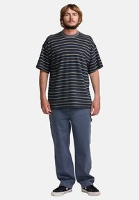 Csíkos, rövid ujjú t-shirt sötétkék alapszínből, világos csíkokkal, laza szabású szürke farmernadrággal és fekete sportcipővel párosítva, látható varrással.