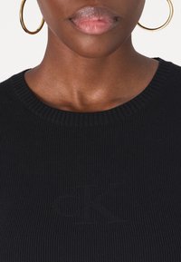 Gros plan d'une personne portant des boucles d'oreilles créoles dorées et un haut côtelé noir avec un logo "ck" subtilement embossé sur la poitrine.