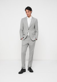 Ανοιχτό γκρι κοστούμι από υφασμάτινο υλικό με υφή· διαθέτει εφαρμοστή σακάκι και παντελόνι με ίσια παντελόνια. Φοριέται με λευκό πουκάμισο και μαύρα παπούτσια.