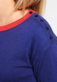 Stickad tröja i djupblått med en röd rund halsringning. Anmärkningsvärda sidoknappar i mörkblått. Mjuk textur, avslappnad design.