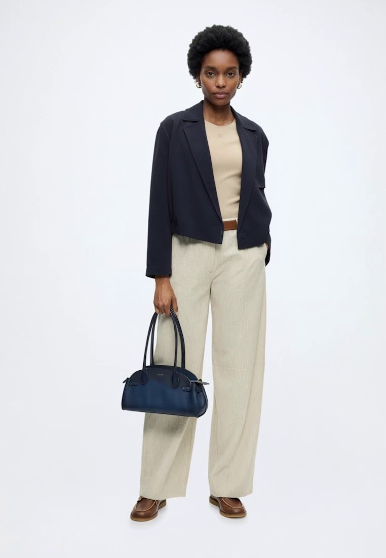 Femme portant un haut beige, un pantalon ample beige clair, un blazer noir court, des chaussures marron, tenant un sac à main bleu foncé avec deux poignées.
