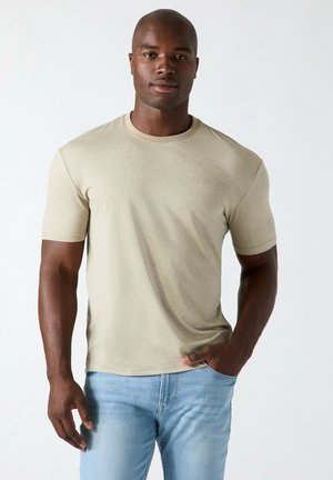 Homme à la peau foncée portant un t-shirt beige à manches courtes et un jean bleu clair, debout avec une main dans la poche devant un fond uni.