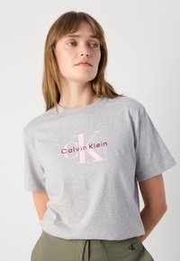 Camiseta de algodón gris con un logo de "Calvin Klein" en rosa y marrón, y un gráfico de "CK", con un corte relajado y mangas cortas.