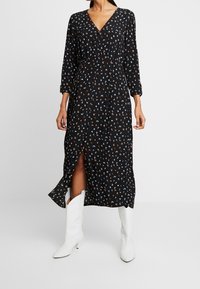 Robe noire à fleurs avec un col en V, des manches longues et une fente à l'avant. Présente un motif floral coloré et est portée avec des bottes de cowboy blanches.