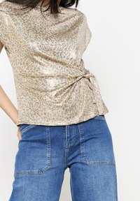 Béžový top s leopardím potiskem a lesklým povrchem, s volným střihem a detaily bočního uzlu, v kombinaci s modrými džínovými kalhotami.