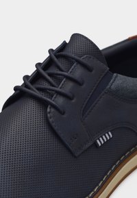 Chaussure à lacets pour homme en bleu marine avec tige texturée, détail en tissu rayé blanc, semelle beige avec surpiqûres blanches.