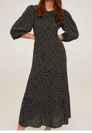 Femme vêtue d'une robe midi noire à pois blancs, manches bouffantes jusqu'au coude et taille cintrée, debout les mains sur les hanches.