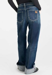 Jeans de mezclilla azul con un corte holgado, que presentan un dobladillo enrollado, un logo naranja contrastante en el bolsillo trasero y detalles de costura visibles.