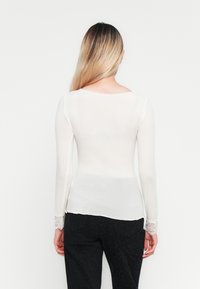 Personne avec de longs cheveux blonds portant un haut blanc ajusté à manches longues avec des poignets en dentelle et un pantalon noir à motifs, tourné de dos sur fond blanc.