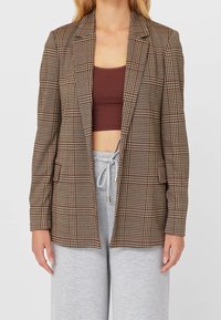 Bruine geruite blazer met pieklapels, enkele knoopsluiting en twee voorzakken. Draagt het over een bruin crop top en grijze joggingbroek.
