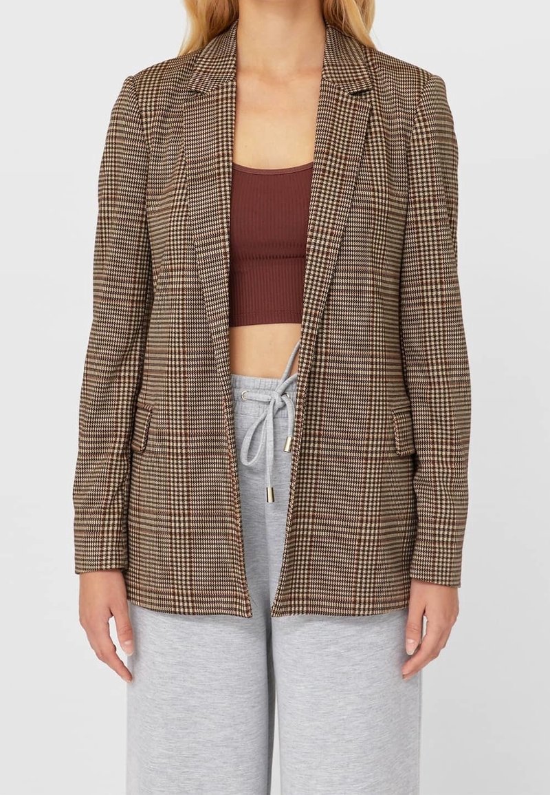 Bruine geruite blazer met pieklapels, enkele knoopsluiting en twee voorzakken. Draagt het over een bruin crop top en grijze joggingbroek.