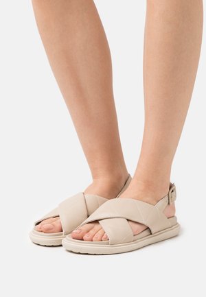 Sandales beiges avec des brides matelassées, présentant un design croisé et une boucle de cheville réglable. Matériau doux et texturé avec une semelle plate.