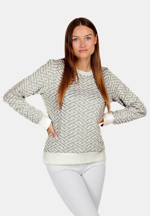 Frau steht mit den Händen in der Hüfte, trägt einen hellgrauen Pullover mit Muster und weiße Hose vor einem schlichten Hintergrund.