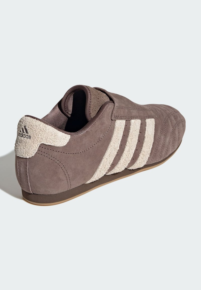 Zapatilla de ante marrón con tres franjas beige, acentos texturizados y un forro suave. Presenta un diseño de perfil bajo y suela de goma.