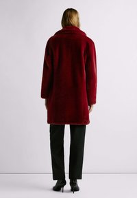 Cappotto rosso in pelliccia sintetica con ampio colletto, maniche a tre quarti e vestibilità comoda, abbinato a pantaloni neri effetto texture e scarpe con tacco alto.