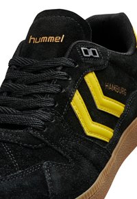 Černé semišové tenisky mají žluté šipkovité akcenty, texturovanou podrážku a na šňůrkách je vyšito logo "hummel" ve zlaté barvě.