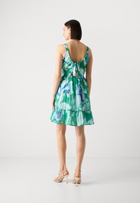 Robe sans manches verte avec des motifs floraux bleus et blancs, design à volants superposés, dos nu avec un nœud, et accompagnée de talons clairs.